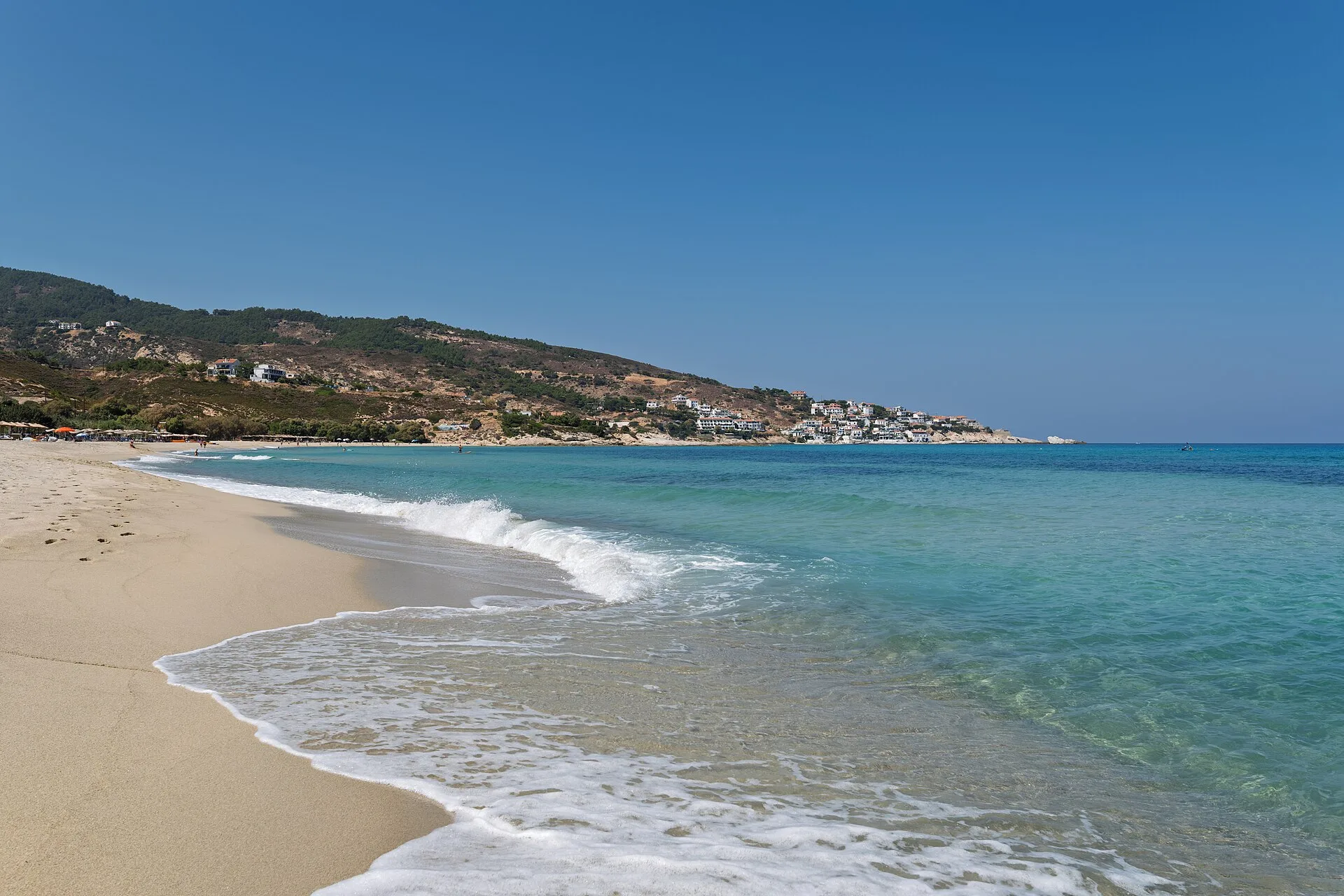 Mesakti Beach auf Ikaria: brechende Wellen über türkisem Wasser, im Hintergrund das Dorf Armenistis am Ende der Bucht